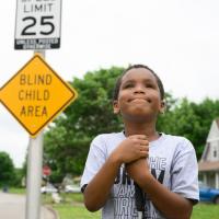 A candid photo of Malakai smiling and holding his hands together as he looks up into the sky. A sign that reads “blind child area” can be seen behind him.