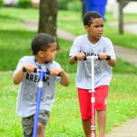 A candid photo of Malakai and his younger brother Kameron riding scooters together outside. Kameron is a young Black boy with short dark hair. He is wearing a short-sleeve gray t-shirt that reads “I am the dream.”