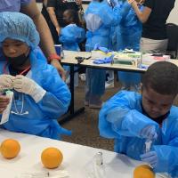 A photo of Malakai and his brother using syringes on oranges while wearing blue surgical clothing.