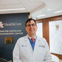 Dr. Marcus Randall, a middle-aged white man with black hair, is standing in the UK HealthCare Radiation Oncology and Radiosurgery Center. He is wearing a white doctor’s coat, a blue sweater, a red tie with white polka dots and spectacles.