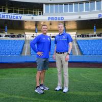 A photo of Matt and UK Baseball Coach Nick Mingione standing on the baseball field. Nick is a white man with short dark hair. He is wearing a blue long-sleeve UK branded shirt with gray shorts and gray Nike shoes.
