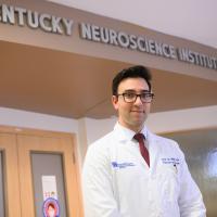 A photo of Dr. Tarek Ali smiling as he stands in front of an office at the Kentucky Neuroscience Institute.