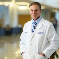 A photo of Dr. Craig van Horne posing in the hallway at UK HealthCare. He is an older white man with short gray hair, wearing a white lab coat over a blue button-up and a blue and white striped tie.
