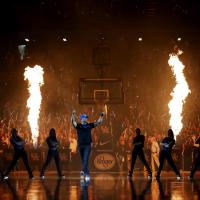 Coach Mitchell dances on the court with the UK dance team dancing behind him and pyrotechnics flaming in the air. The stadium is packed with people.