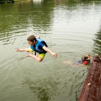Max, wearing a life jacket, strikes a pose as he jumps off the dock into the lake. His younger brother, also wearing a life jacket, is visible in the lake at the end of the dock.