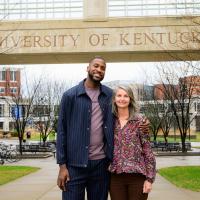 A photo of Michael and Meg Shake smiling for the camera with their arms around each other while on the University of Kentucky campus. Meg is an older white woman with mid-length gray hair. She is wearing a multicolor long-sleeve paisley top and black slacks.