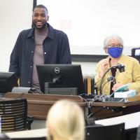 A candid photo of Michael and a UK professor standing at the front of a classroom before giving his presentation. She is an older white woman with short gray hair. She is wearing a yellow denim jacket over a black shirt and gold necklaces.