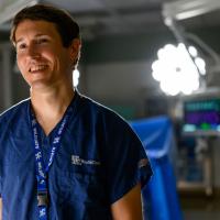 A portrait of Dr. James, a young-looking man with brown hair, dressed in blue scrubs, standing in a surgery room.