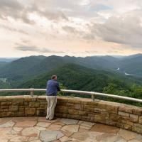 A landscape photo of Michael with his back facing the camera as he stands on a gated ledge that looks out over mountains and trees.