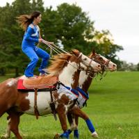 Miko stands atop two running horses, with a foot on each horse’s saddle. They are running in a field.