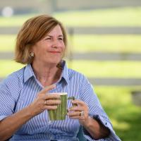 A photo of Elizabeth (E) Barr looking off-camera as she sits outside with a cup of coffee. E is an older white woman with short brown hair. She is wearing a long-sleeve blue-and-white-striped shirt.