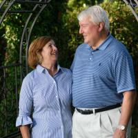 A candid photo of E and her husband smiling at each other while standing under a metal gateway in their garden. Her husband is an older white man with white hair. He is wearing a blue short-sleeve polo that is tucked into his khaki pants.