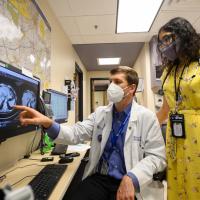 A photo of Dr. Michael Cavnar pointing at a patient’s scan on a computer screen as Dr. Reema Patel looks on. Dr. Cavnar is a white man with short brown hair. He is wearing a white lab coat over a long-sleeve blue button-up shirt. Dr. Patel is a young South Asian woman with long curly black hair. She is wearing a yellow floral print dress, a gray face mask, a pair of dark-rimmed glasses, and a stethoscope around her neck.