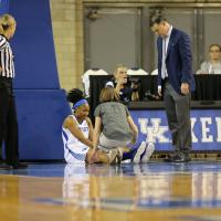 Ogechi sits at the edge of the court, surrounded by Coach Matthew Mitchell, the team physician, and a referee. Ogechi is holding her knee and has her legs straight out in front of her.
