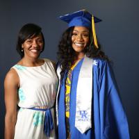 Ogechi poses for a photo in her graduation cap and gown, a yellow dress under her robes, and a silver UK Athlete stole. She is smiling broadly and her hair is loose and curled. Standing next to her is Tiffany Hayden, former Women’s Basketball academic advisor and current Assistant Athletic Director for Diversity, Equity and Inclusion. Tiffany, a young-looking Black woman, is wearing a white dress with floral print and is smiling.