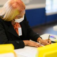 Ogechi, dressed in a black blazer and orange blouse, works at a desk at the UK HealthCare Sports Medicine clinic. Her hair is long, straight, and blonde.