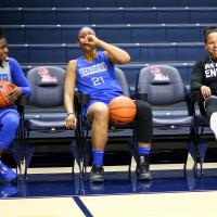 Ogechi sits on the sidelines, wearing a knee brace and a UK jersey. She is laughing and holding a hand up to her mouth. Coach Amber Smith is sitting to one side of her and is also laughing. Another teammate is sitting on the other side of Ogechi and is grinning at both of them.
