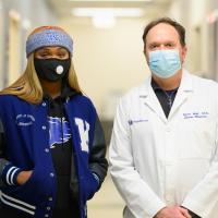 Ogechi and Dr. Mair pose for a photo together inside the Sports Medicine clinic. Both are wearing masks. Ogechi is wearing a blue Kentucky basketball jacket, a black and blue Wildcat hoodie, and a gray UK headband. Dr. Mair is wearing a white lab coat.