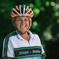 Patty smiles brightly for a photo while wearing an orange and silver biking helmet and white and green cycling kit.