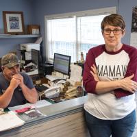 Debbie leans against the Gold City Towing office reception desk while Chuck sits in the chair behind her and smiles.