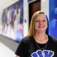 A photo of Julie smiling for the camera in one of the hallways of the hospital. Decals of UK basketball players can be seen in the background on the wall.