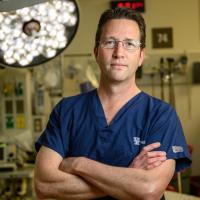Dr. Andrew Bernard, a white adult man with brown hair, stands confidently in an empty operating room with his arms crossed. He is wearing a blue set of scrubs, and a pair of glasses.