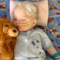 Rory holds his stuffed lion toy as he sleeps. He is wearing a blue striped shirt and has tubes in his nose and taped to his head.
