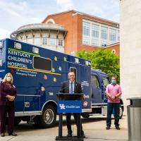 Standing behind a podium outside of the hospital in front of a blue Kentucky Children’s Hospital ambulance—Dr. Day prepares to speak to his colleagues and the community. He is dressed in a black suit with a blue shirt and blue tie.