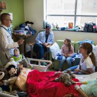 Dr. Day stands in a young patient’s room speaking to their family—the room is filled with posters, drawings, toys and stuffed animals.