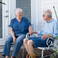 A candid photo of Carol Bailey and her husband Rick Bailey smiling and holding hands as they sit on the porch outside of their home. Carol, who is Rick’s caretaker, is an elderly white woman with short white hair. She is wearing a short-sleeve blue shirt with blue jeans. Rick is an elderly white man with short graying hair. He is wearing light-sleeve light blue button-up with jean shorts.