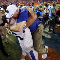 A UK football player lifts Coach Stoops off the ground in a huge hug on the sidelines of a football game.