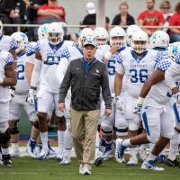 Coach stoops, wearing a gray jacket, leads a team of Kentucky football players onto the field. He has a whistle in his mouth and is wearing a UK baseball cap.