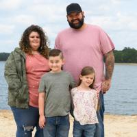 Taytum and her family—including her parents and brother, Silas—pose for a group portrait at the edge of a lake. Her parents are both larger—her mom, who has curly brown hair, is wearing a pink top and camo jacket, and her dad, who is bearded and has a tattoo sleeve, is wearing a pink tee. Silas is taller than Taytum and has his arm around her. Everyone is smiling.
