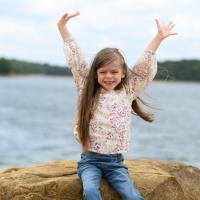 Taytum sits on a rock next to the lake. She holds her hands up in the sky and grins.