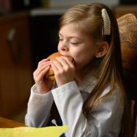 Taytum chomps down on a McDonald’s cheeseburger, held in both hands. She is still wearing her white coat and scrubs.