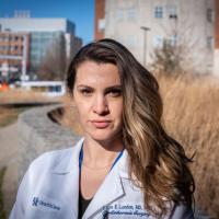 A portrait of Dr. London-Bounds, standing outside the hospital in a white coat and a blue shirt. Her hair is loose and pushed to one side, and she is wearing a small gold chain and green hoop earrings.