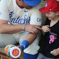 T.J. sits in the dugout with a young fan wearing aa baseball cap. T.J. is holding a JoyJar—a brightly colored plastic jar filled with toys—and is pointing at the kid playfully. Both are smiling.