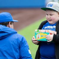 T.J. crouches down to talk to a young kid wearing a baseball hat. The kid is holding a brightly-colored box. T.J. is wearing a baseball cap and a blue jacket.