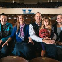 Todd, his wife, and their three kids pose for a family photo with a basketball in UK gymnasium seats.