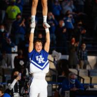 A female cheerleader stands on Travis’ hands during a cheer routine.There are two other cheerleading pairs in the same formation on both sides of him.