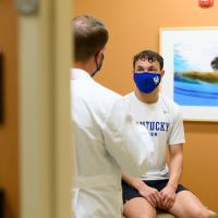 An over-the-shoulder shot of Travis having a conversation with Dr. Scott Mair. Dr. Mair is a white man with light brown wavy hair. He is wearing a white lab coat over blue scrubs, and a face mask.