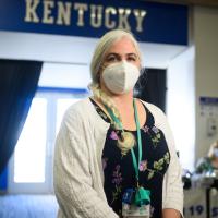 Karla Bertagnole, a middle-aged woman with long white hair in a single braid, stands inside the Kroger Field stadium. She is wearing a white facemask, a navy dress with floral designs, and a white cardigan.