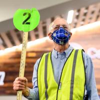 Bob Evans, an older man with grey hair, stands inside the Kroger Field stadium directing people. He is wearing a blue facemask, a green reflective vest, a blue shirt, and is and holding up a number 2 sign.