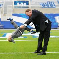 Coach Stoops, wearing a suit, holds his son’s hands as his son flips through Coach’s arms. His son is upside down, mid-flip, and is wearing a gray sweatshirt gray pants and blue sneakers.