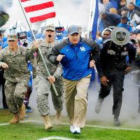 Coach Stoops runs out onto the field ahead of two National Guardsmen in uniform, one holding a flag, as well as another uniformed person. The football team is behind them.