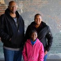 A photo of Whitley, her father, Vincent Smith, and Essence standing together and smiling for the camera. Vincent is an older African-American man with a bald head, graying goatee, and glasses. He is wearing a black hoodie over a gray t-shirt and blue jeans.