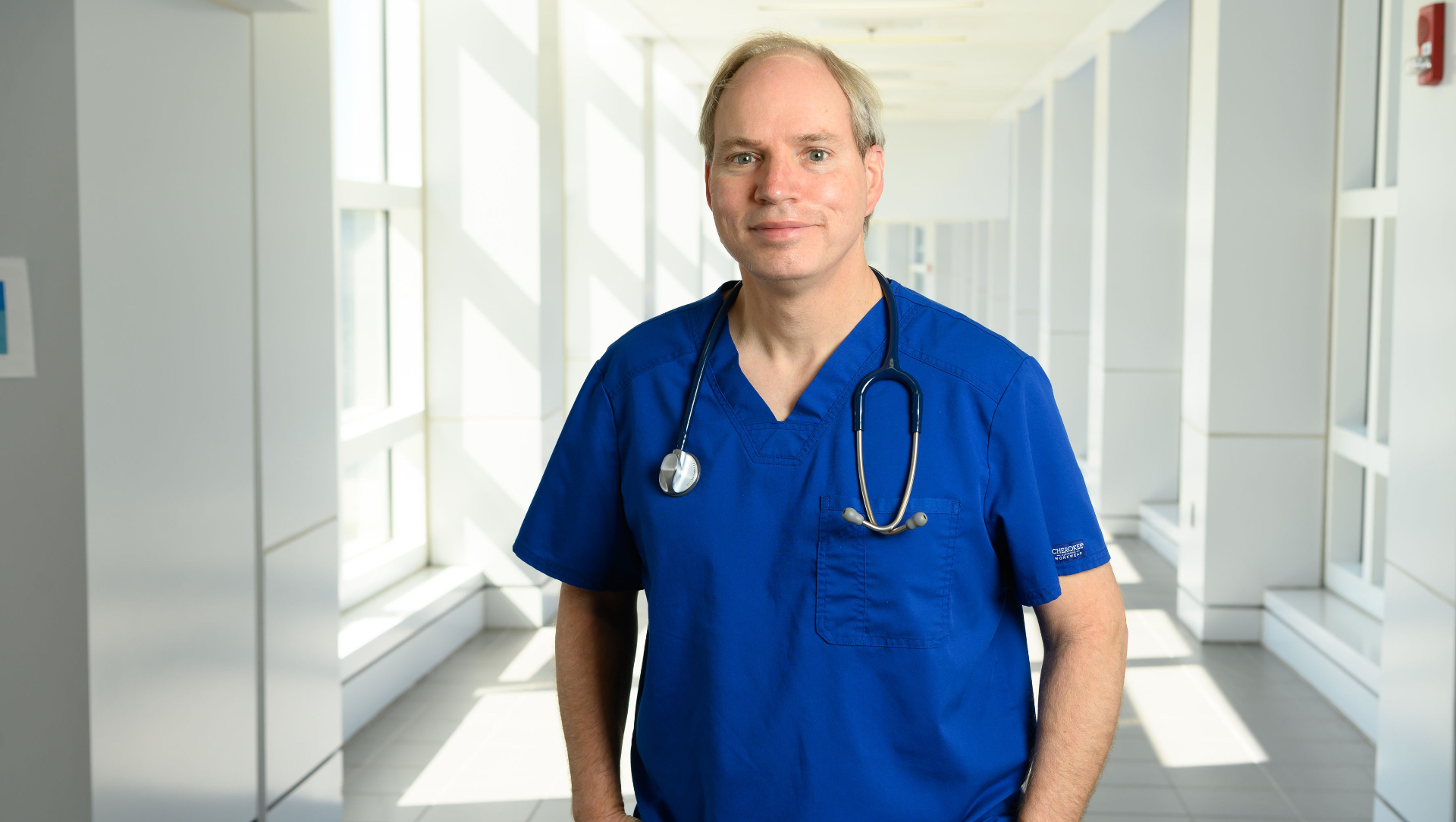 Dr. Michael Anstead, a middle-aged white man with grey hair, stands in the hallway of UK HealthCare wearing blue scrubs with a stethoscope around his neck.