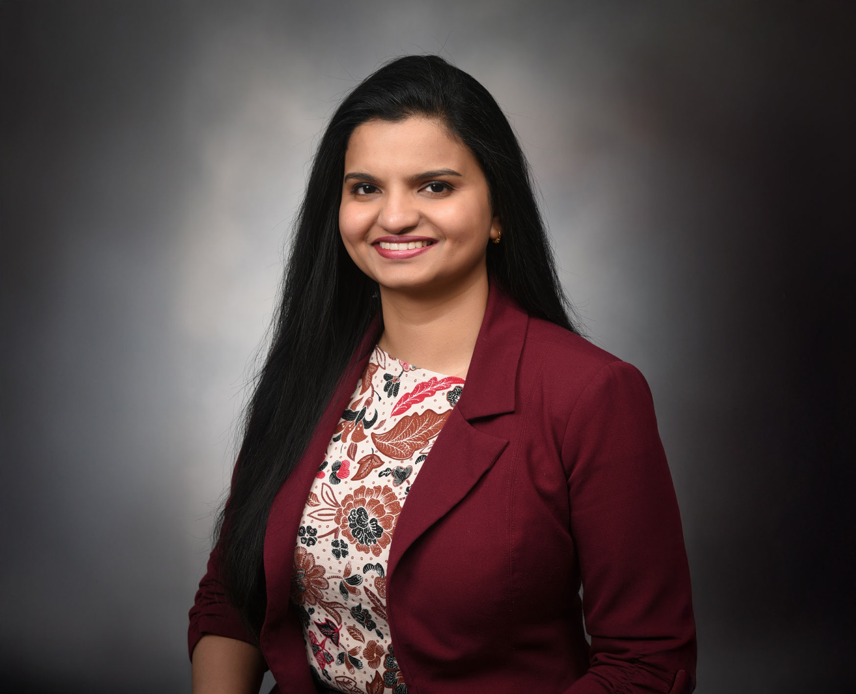 Dr. Sonu Abraham, seated for a portrait. She's smiling and wearing a floral shirt with a red jacket over it.