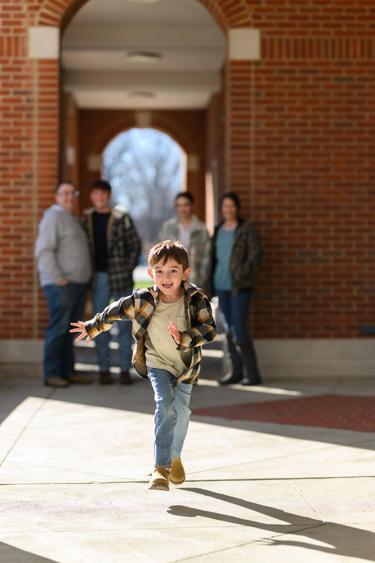 Granger Smith, very prominent in the foreground, runs toward the camera lens offscreen. His family is visible, though out of focus, in the background near a brick building.