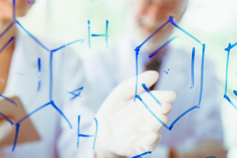 A gloved hand writes chemical structures on a transparent surface in a laboratory setting.